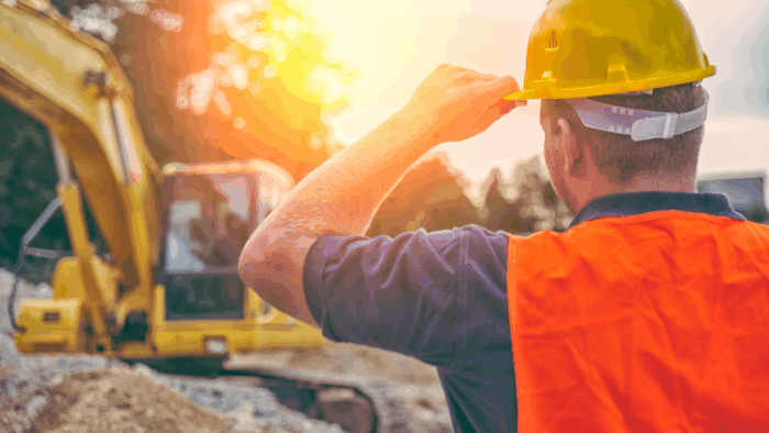 A construction worker holding a yellow safety helmet, standing and observing an active construction site with building materials and machinery in the background.