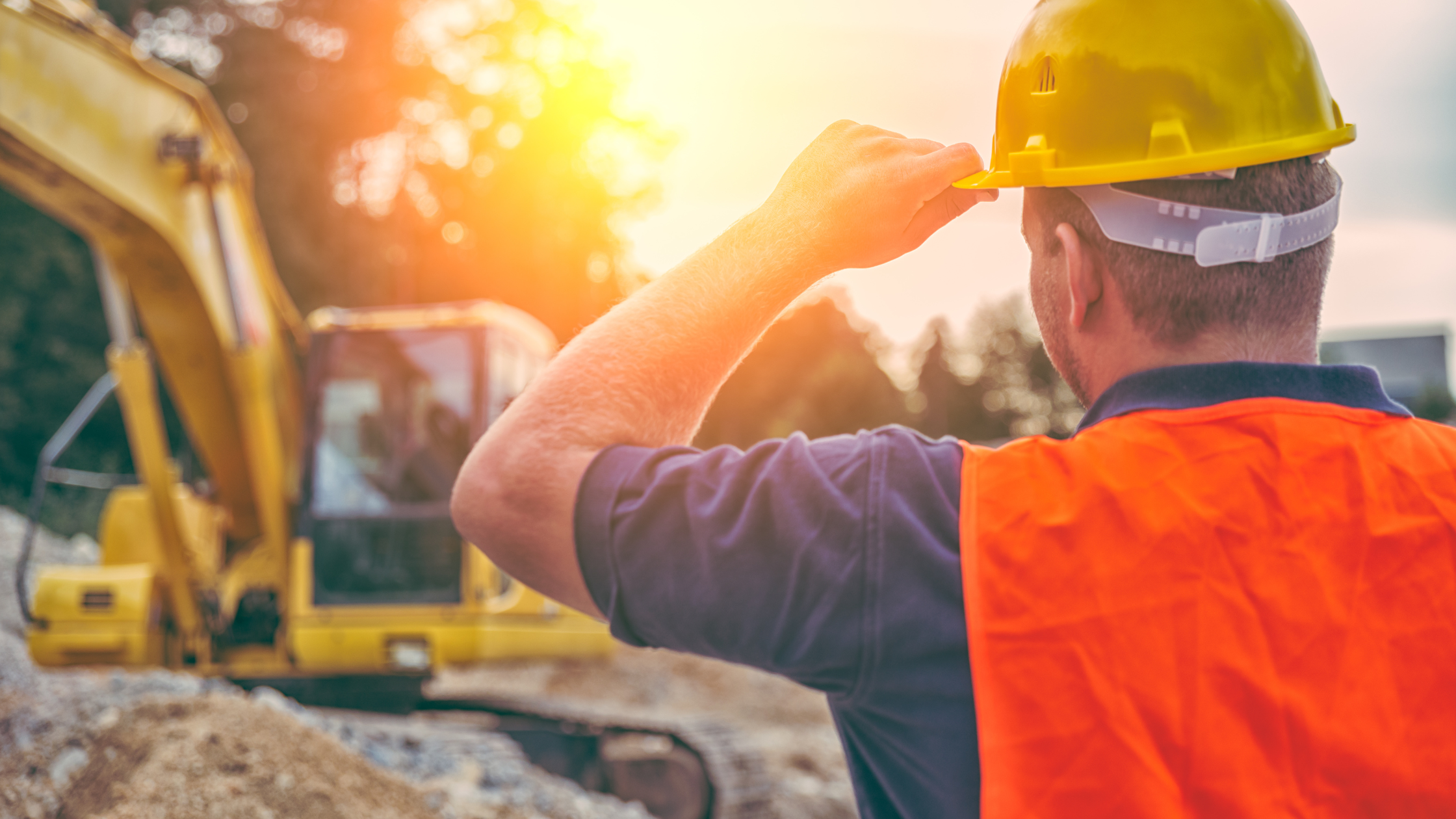 A construction worker holding a yellow safety helmet, standing and observing an active construction site with building materials and machinery in the background.